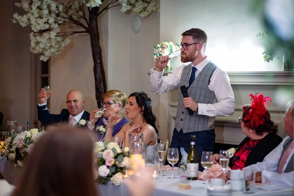 A groom stands and gives his wedding speech while guests seated at the top table raise their glasses for a toast.