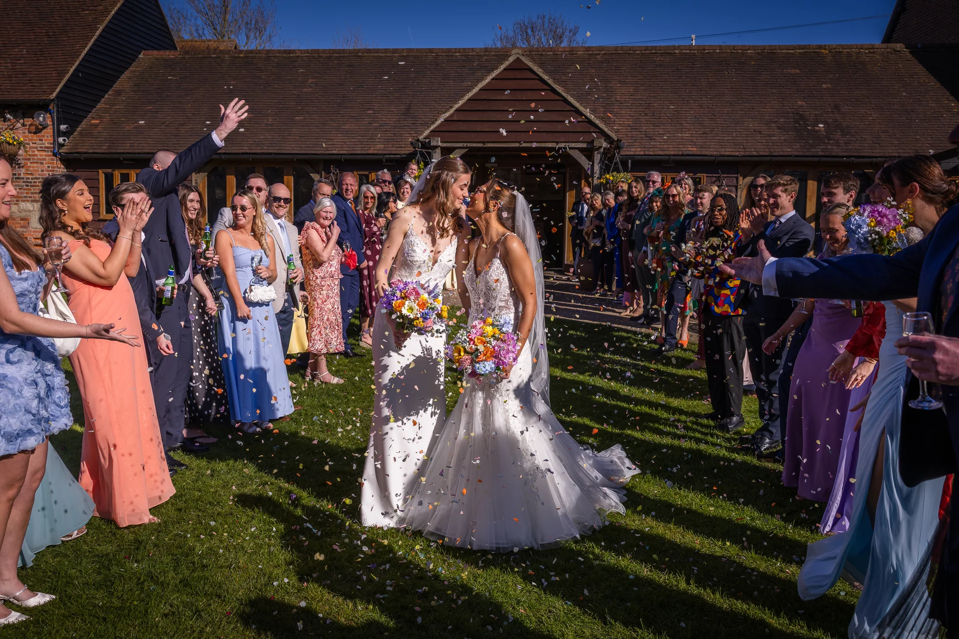 Two brides share a tender kiss as guests shower them with colorful confetti on a sunny day outside a rustic barn venue.