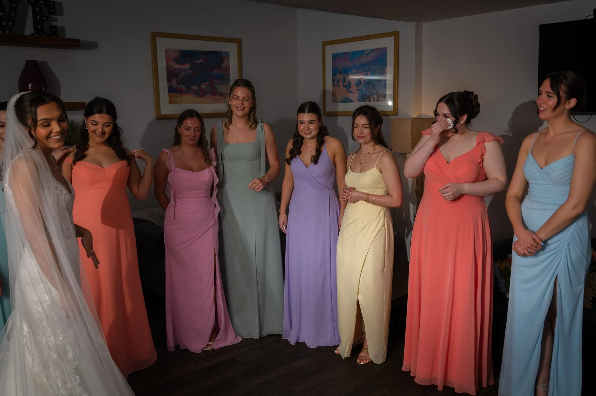 A beautiful bride reveals her wedding dress to an emotional group of bridesmaids wearing colourful pastel dresses during morning preparations.
