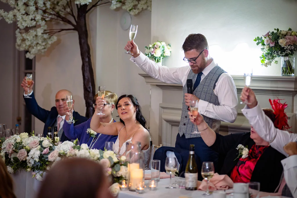 A groom delivers a speech while standing at the top table, raising a glass alongside the bride and guests during the wedding breakfast.