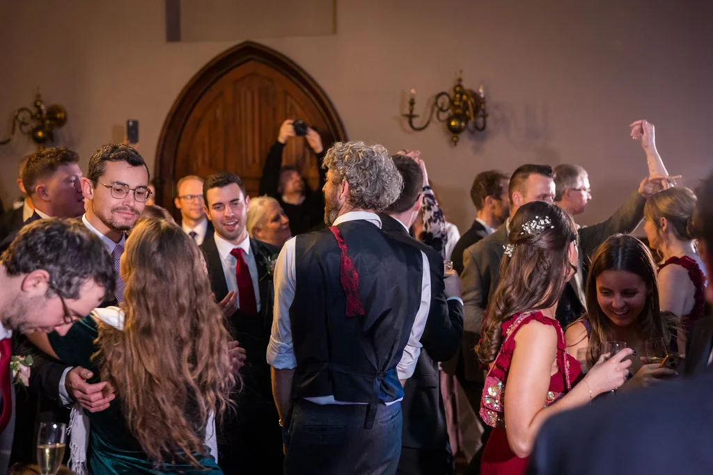 A lively candid shot of wedding guests mingling and celebrating during the evening reception inside a venue with a stone archway in the background.