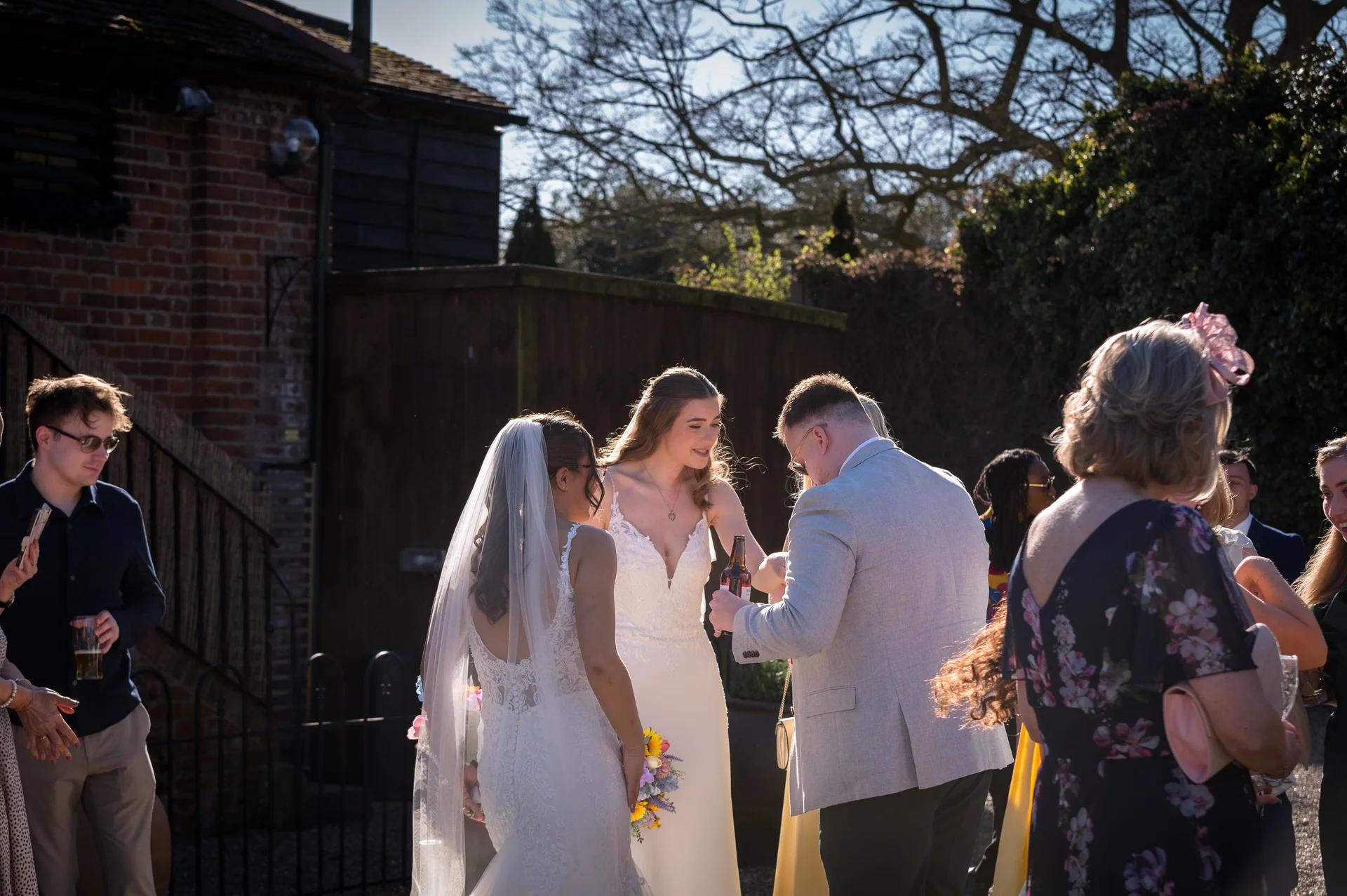 Wedding guests mingle in a sunlit courtyard outside a rustic barn while a bride and groom share a moment during the celebrations.