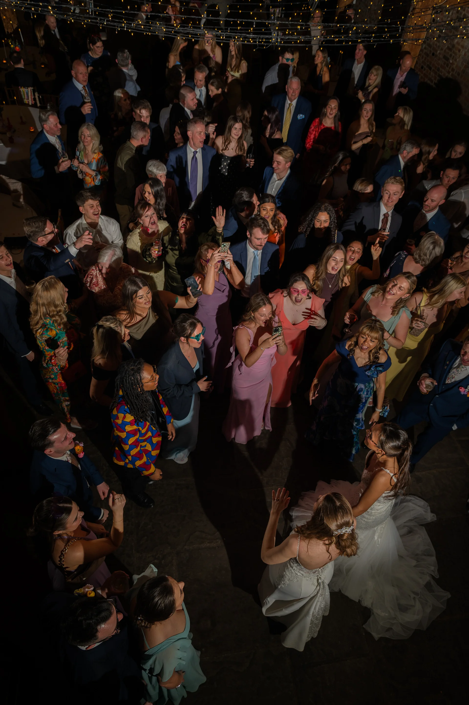 An elevated overhead view captures a lively wedding reception dance floor filled with enthusiastic guests dancing around the newlyweds in a dimly lit venue draped with string lights.