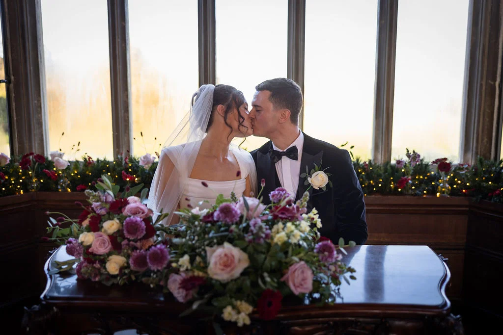 Newlywed bride and groom share a tender kiss seated at a wooden table adorned with rich plum and blush floral arrangements in front of large panelled windows.