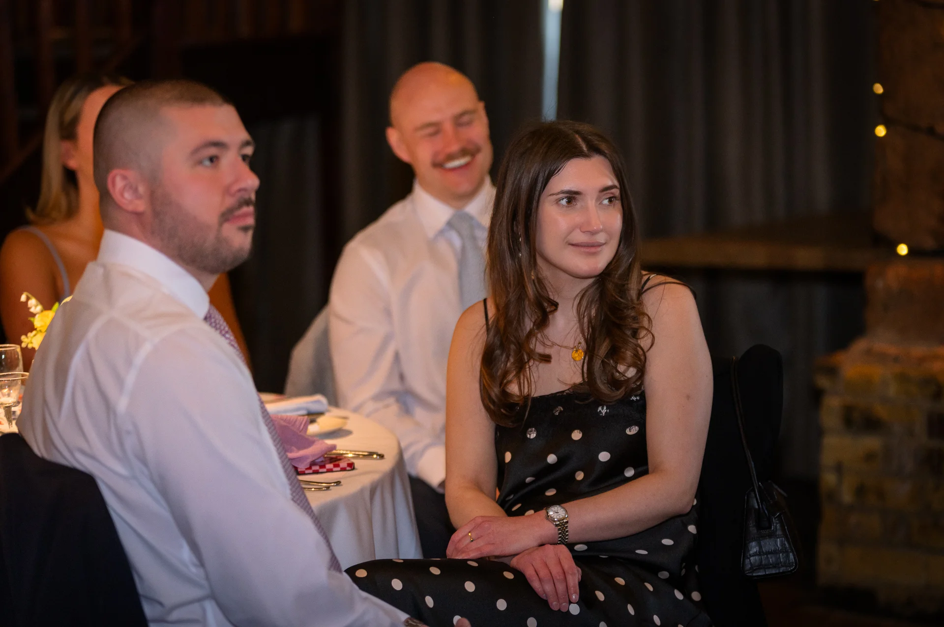 Wedding guests seated around a table listening attentively and smiling during the reception speeches.