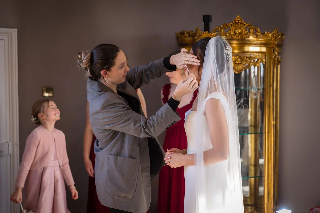 A bride receiving final touch-ups to her makeup during bridal preparations, wearing a sleek white gown and chapel veil as a flower girl and bridesmaids look on.