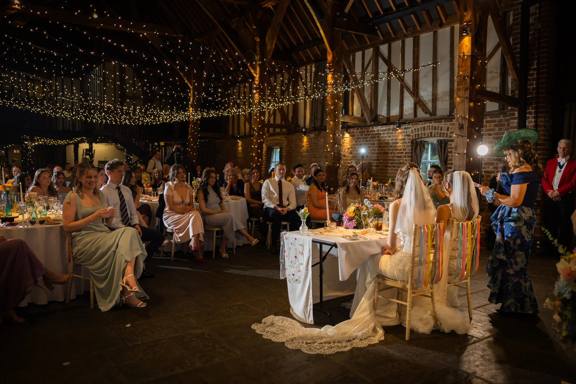Two brides seated at a top table during wedding speeches inside a rustic oak-framed barn decorated with an extensive fairy light canopy and exposed brick walls.