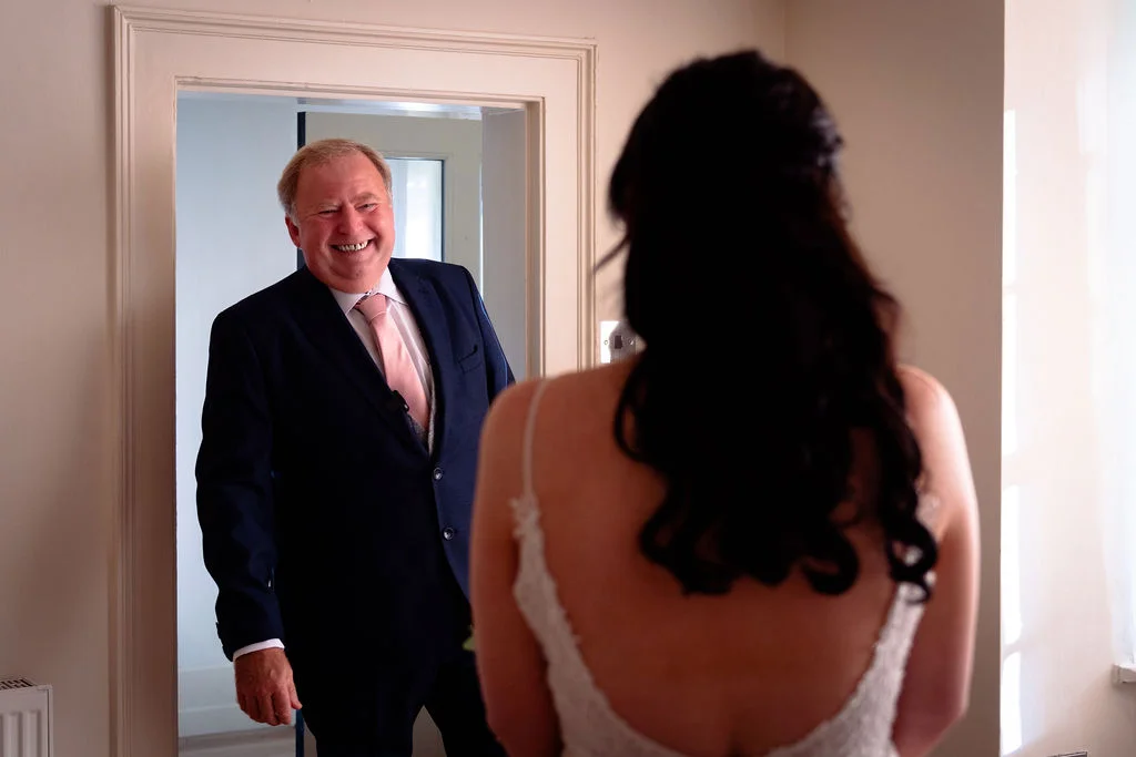 A smiling father of the bride reacts with pure joy during an emotional first look in a doorway as his daughter looks on from behind.