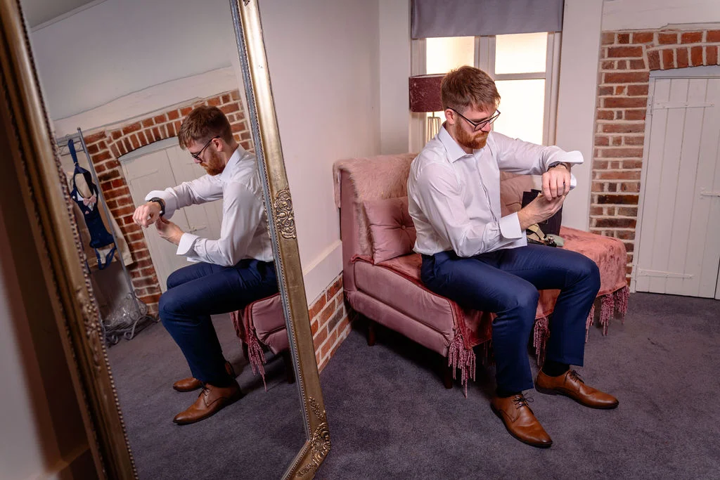 A groom in a white shirt and blue trousers sits on a pink velvet chaise longue while adjusting his cuffs, reflected in a large ornate mirror.