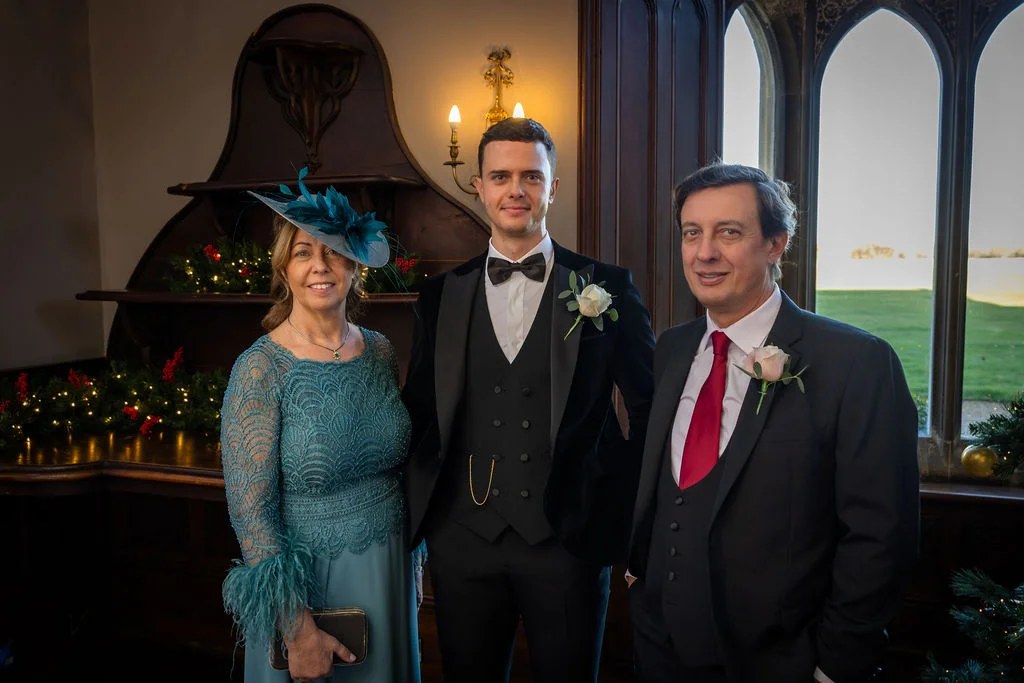 A formal family group portrait featuring a groom in a black tuxedo flanked by his parents standing indoors near arched windows.