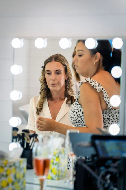 A bride with curled hair and hair clips looks intently in a lighted vanity mirror while getting ready on her wedding morning.