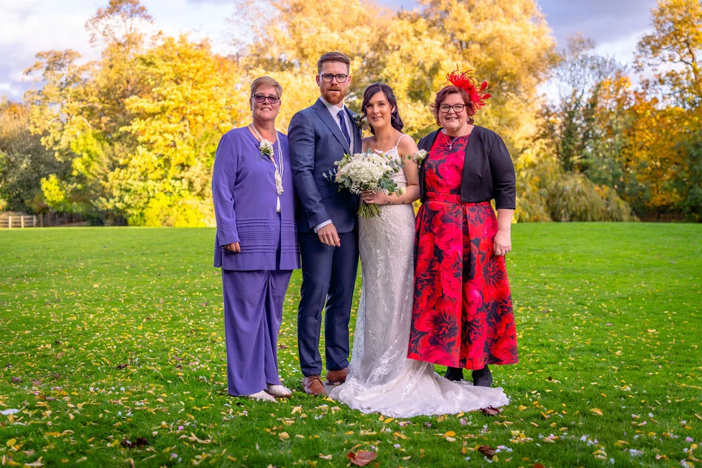 A bridal party group stands on a lush green lawn scattered with fallen autumn leaves against a backdrop of vibrant yellow and golden trees.