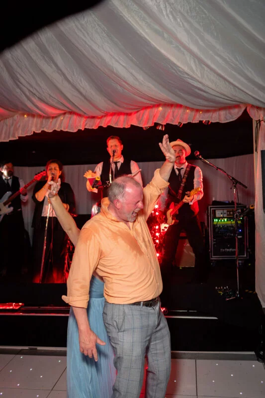 A guest dances enthusiastically with arms raised on a lit dance floor inside a marquee during a lively wedding reception.