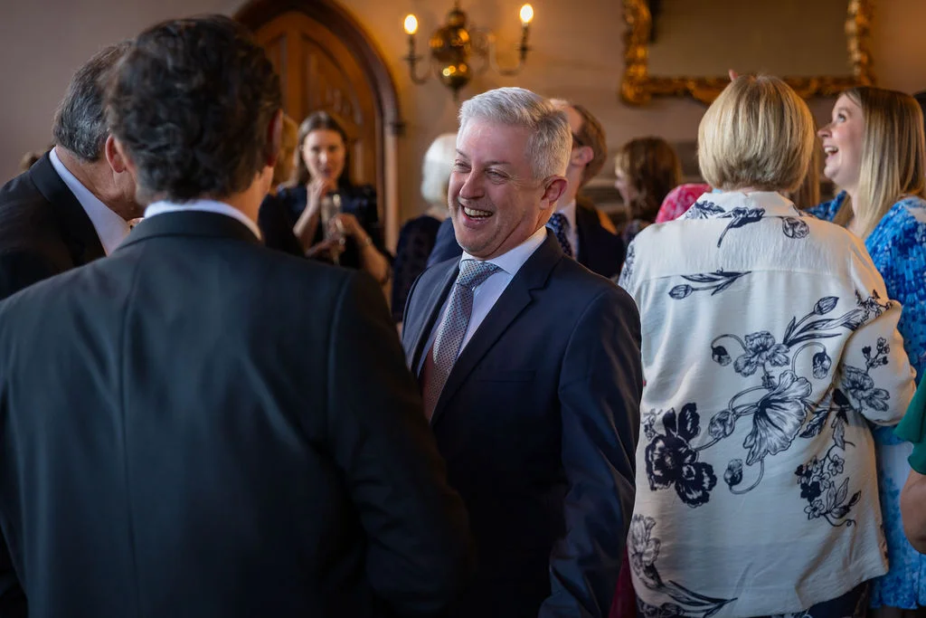 A smiling guest in a dark suit and patterned tie laughs warmly while mingling during the wedding reception drinks.