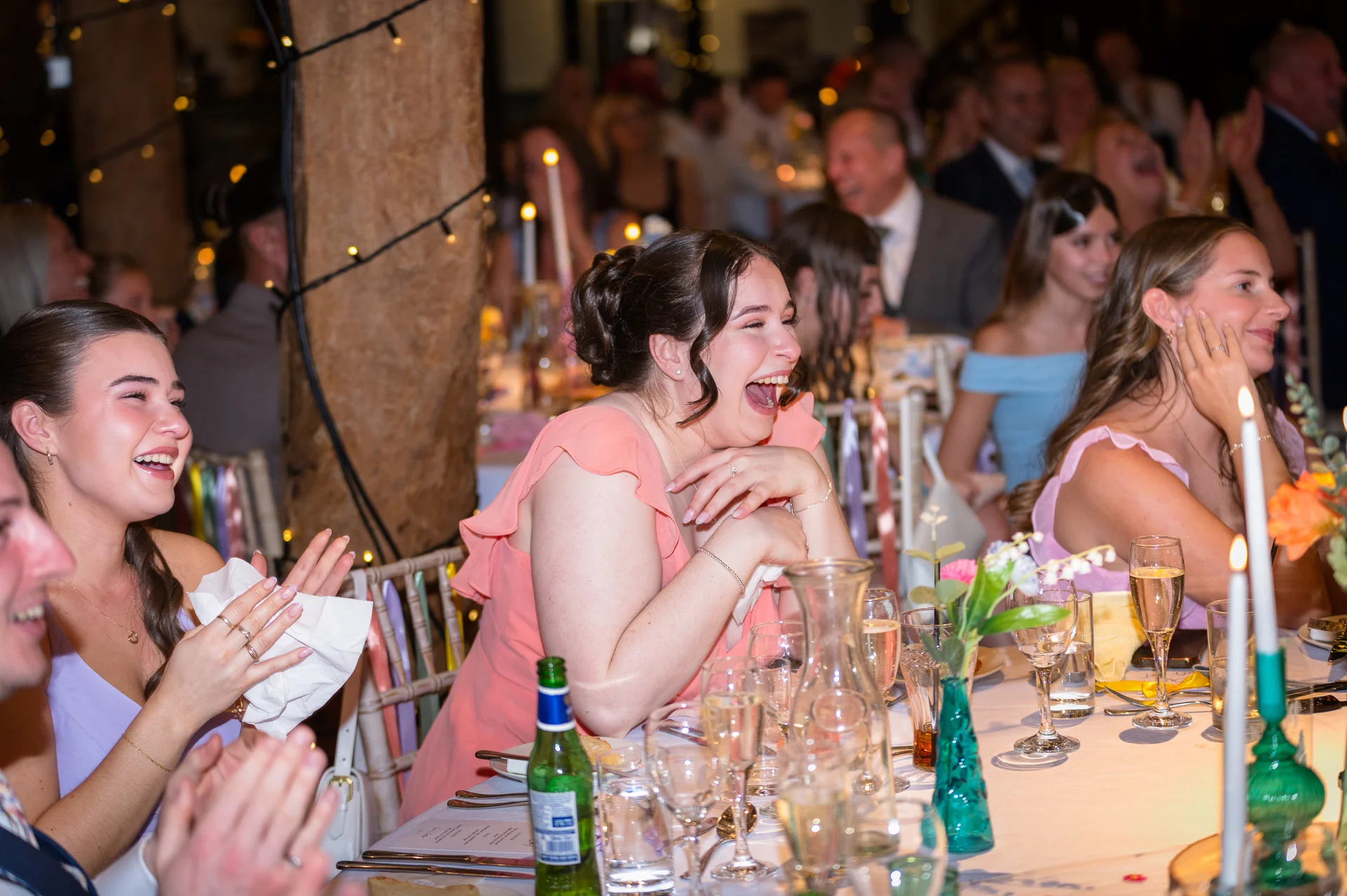 Wedding guests laugh uncontrollably during the speeches at a candlelit barn reception, seated at a long banquet table adorned with glassware and tapers.