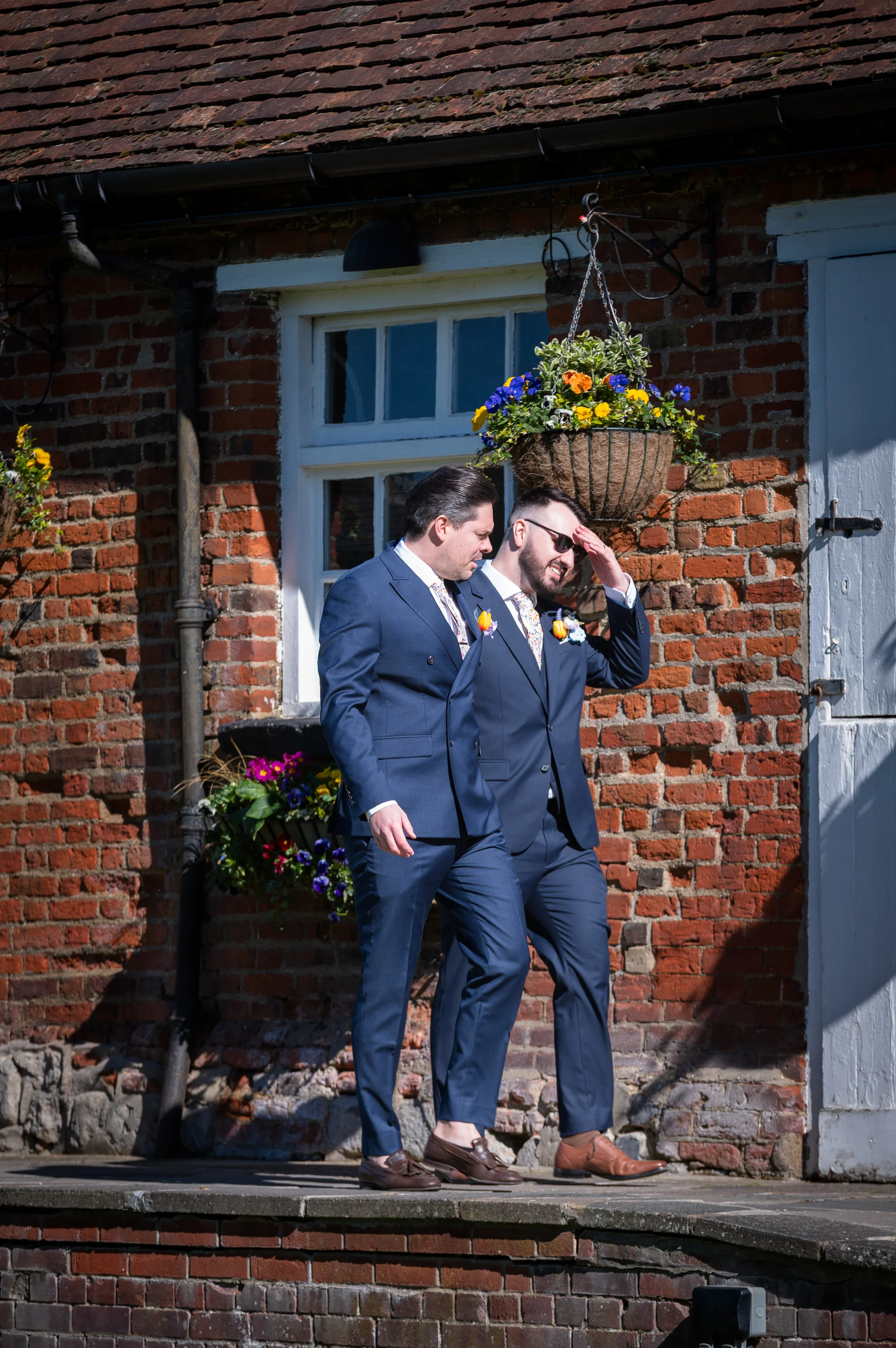 Two grooms in matching navy suits walk together outdoors in bright sunshine along a weathered red brick building with hanging baskets.
