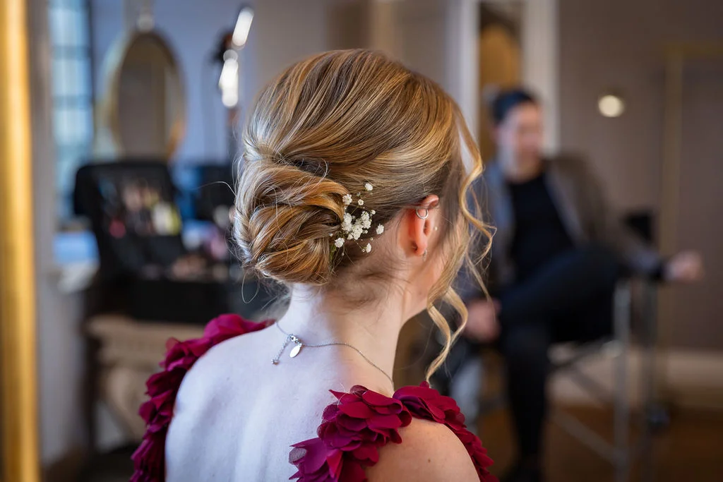 A close-up back view of a bride showcasing an intricate textured bun adorned with delicate baby's breath flowers during her morning preparations.