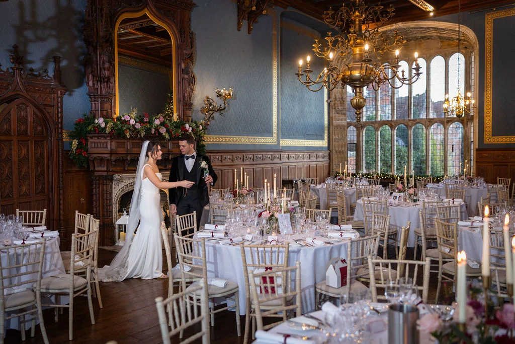 Newlyweds stand inside a grand panelled ballroom featuring towering windows, large crystal chandeliers, and neatly set round tables for a wedding breakfast.