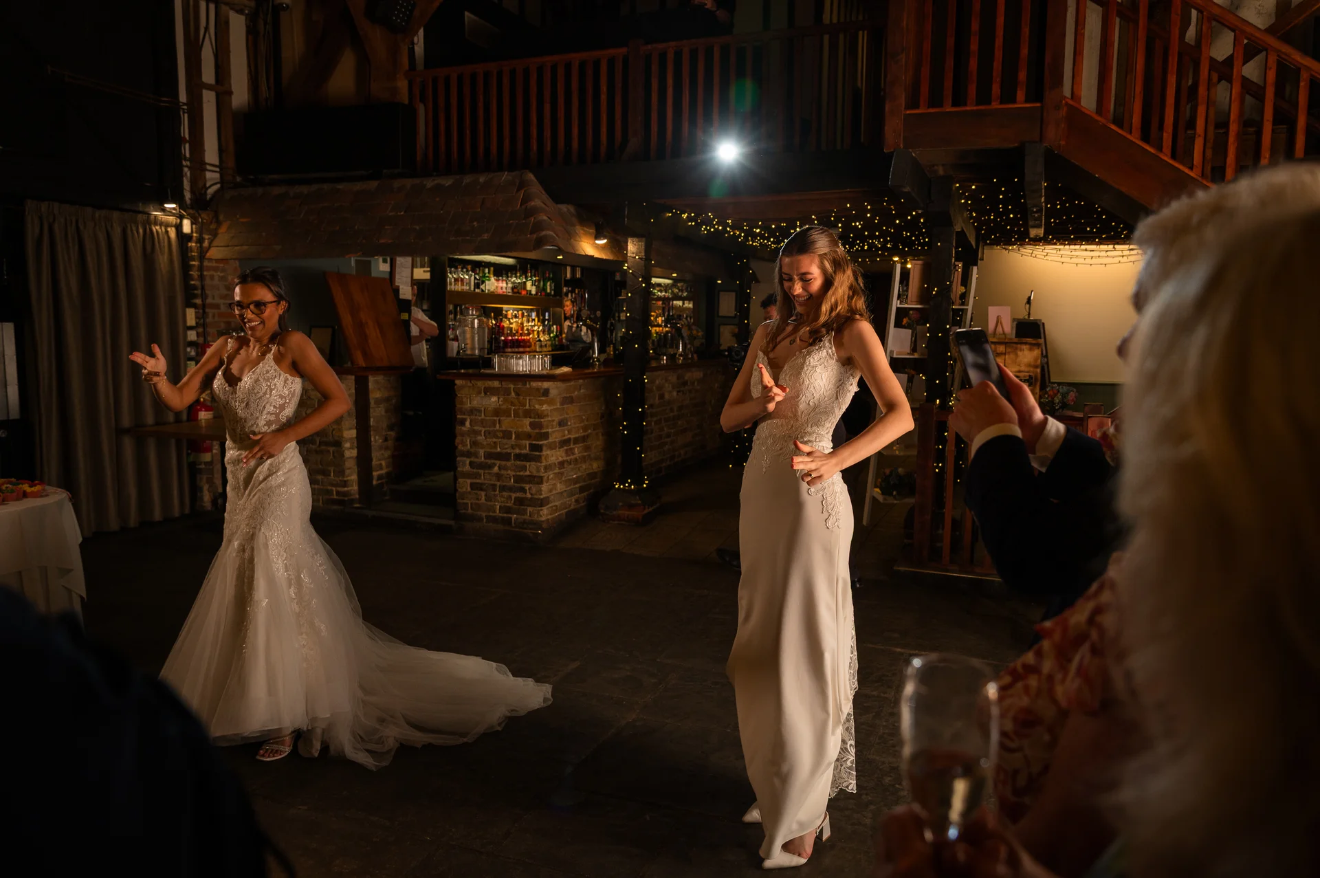 Two brides are joyfully dancing together in a rustic barn venue while guests record the moment on their phones during the evening reception.