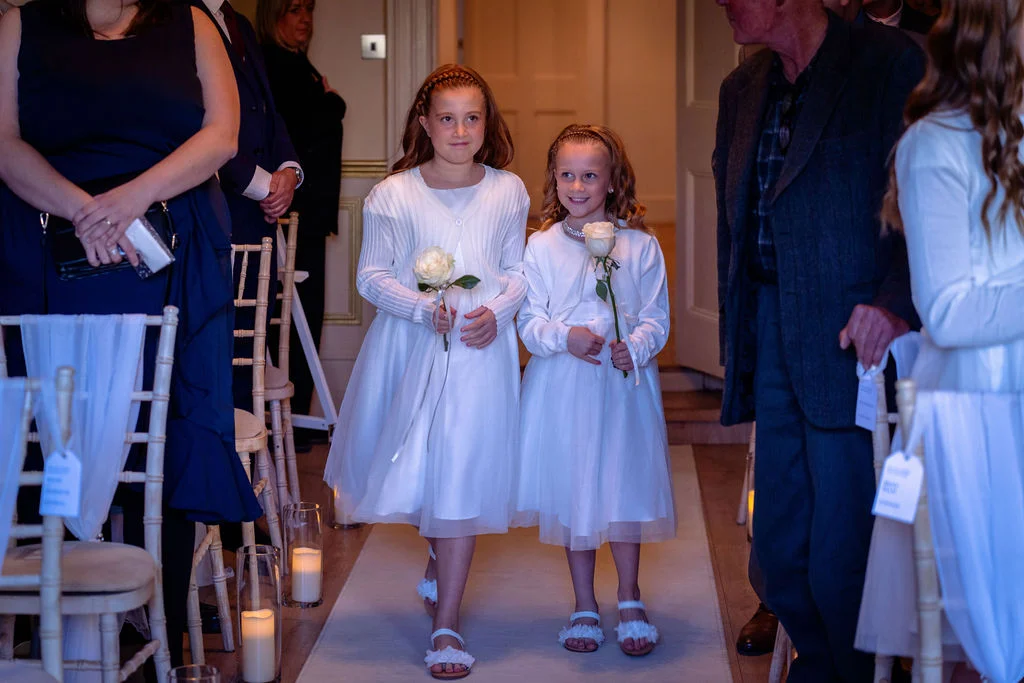 Two young flower girls in white dresses walk down the aisle carrying single white roses during a wedding ceremony.