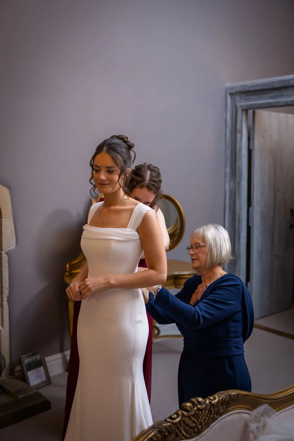 A bride in a sleek crepe gown with a square neckline has the finishing touches applied by her mother during the morning bridal preparations.