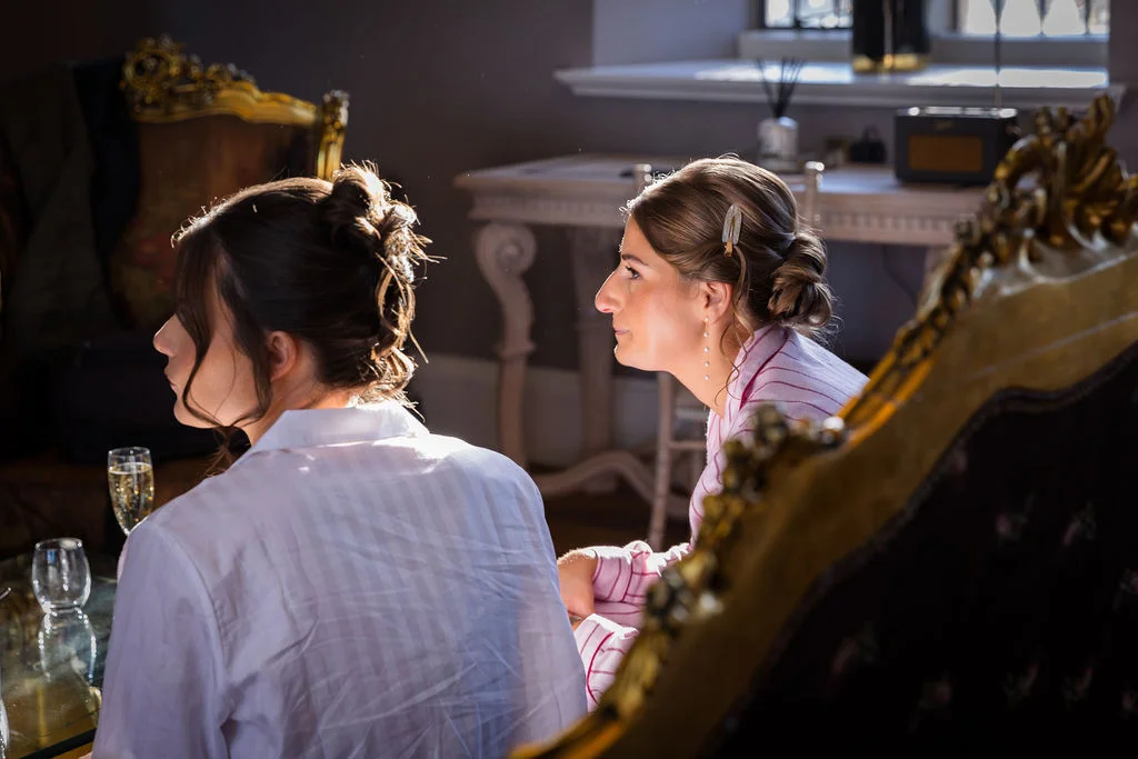 Two bridesmaids relax in matching pyjamas with champagne while getting ready in a sunlit room before the wedding ceremony.
