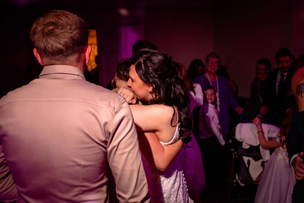 A bride and groom share an intimate embrace on a dimly lit dance floor surrounded by wedding guests during the evening reception.