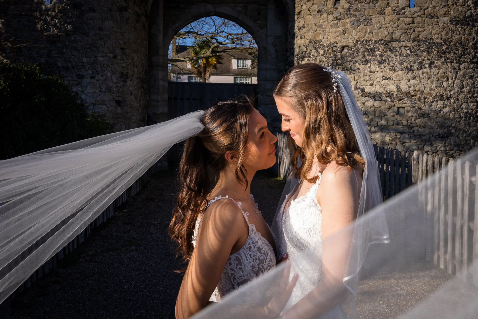 Two brides in lace wedding gowns gaze lovingly at each other outdoors with a dramatic veil blowing in the wind against a historic stone castle wall.