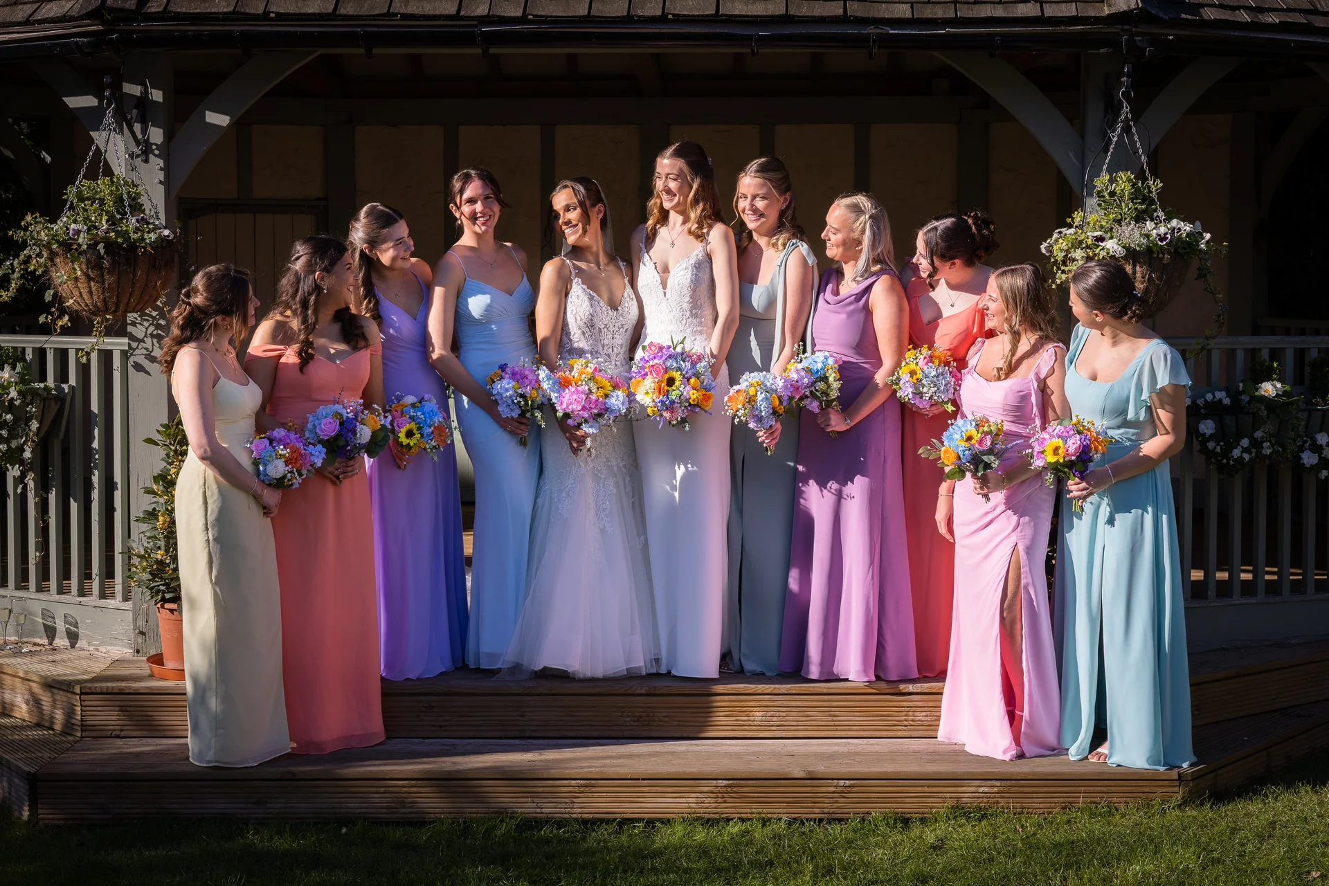 Two brides and a large group of bridesmaids in colourful dresses pose together outdoors in front of a wooden gazebo on a sunny day.