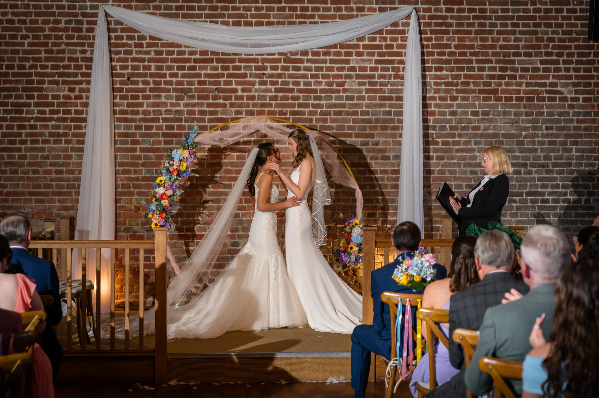 Two brides stand face-to-face during their wedding ceremony in a rustic room with an exposed brick wall, floral circular arch, and white fabric draping.