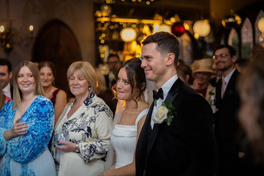 The bride and groom stand together amidst guests during an evening celebration, with warm fairy lights glowing in the background.