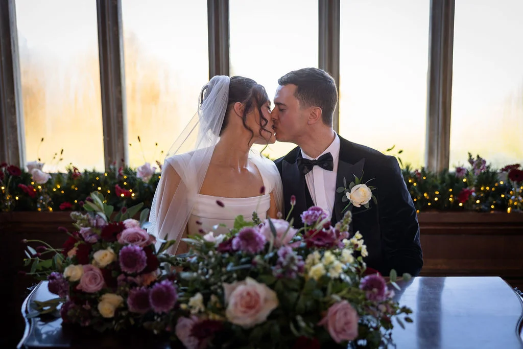 Newlywed bride and groom share a tender kiss in front of a large window with rich floral centrepieces and fairy light decor.