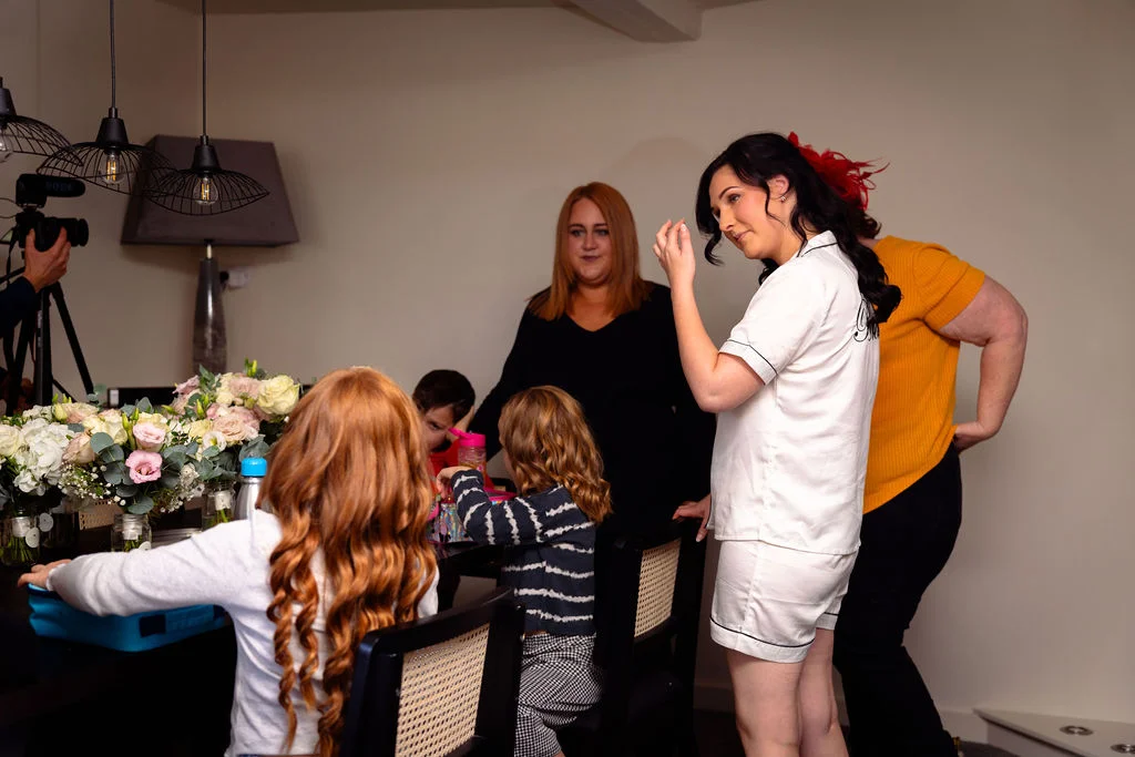A bride in matching white pyjamas gets her hair and makeup styled indoors while surrounded by family members and children sitting at a table decorated with floral arrangements.