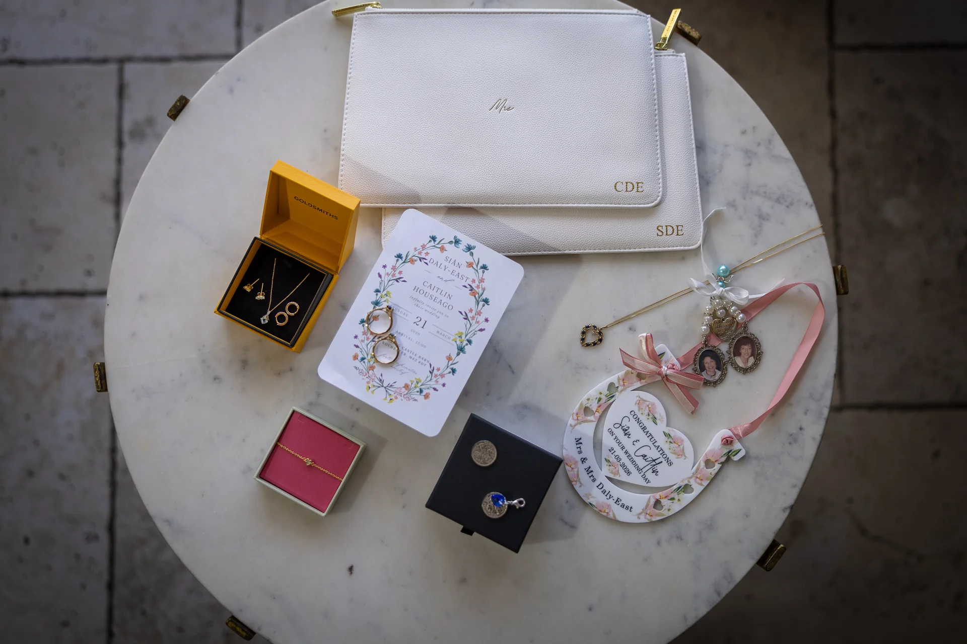 An elegant bridal flat lay arranged on a round marble table showcases wedding stationery, personalised white leather pouches, gold jewellery in boxes, and a floral horseshoe charm.