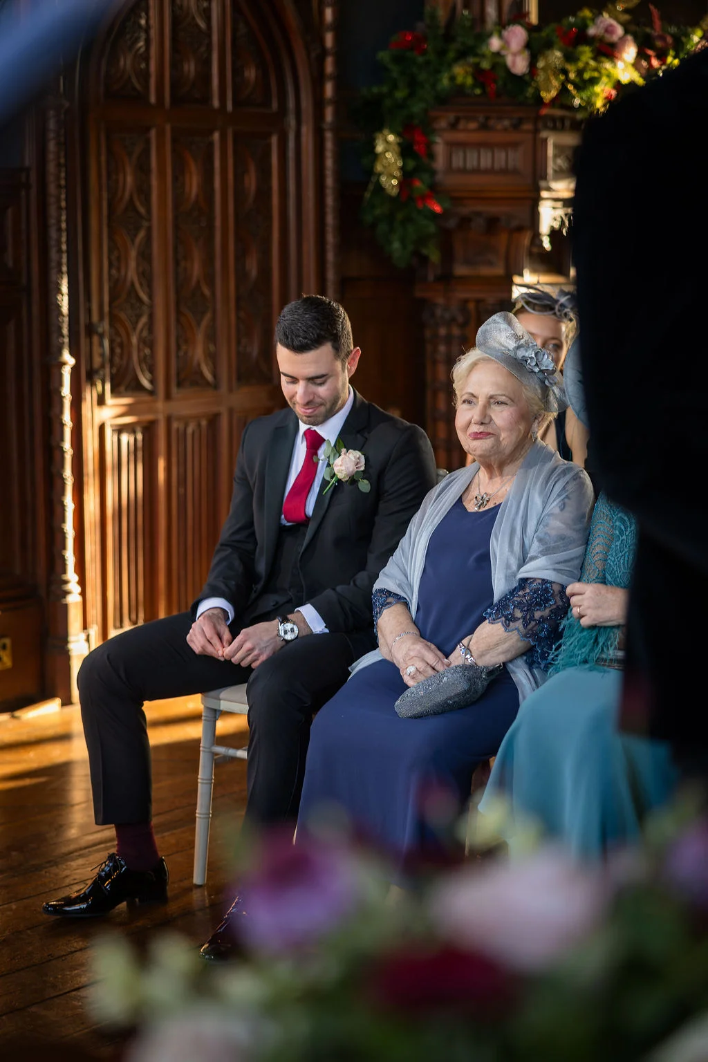 A groom in a black suit with a red tie and an elderly guest wearing a fascinator sit together inside a wood-panelled room.