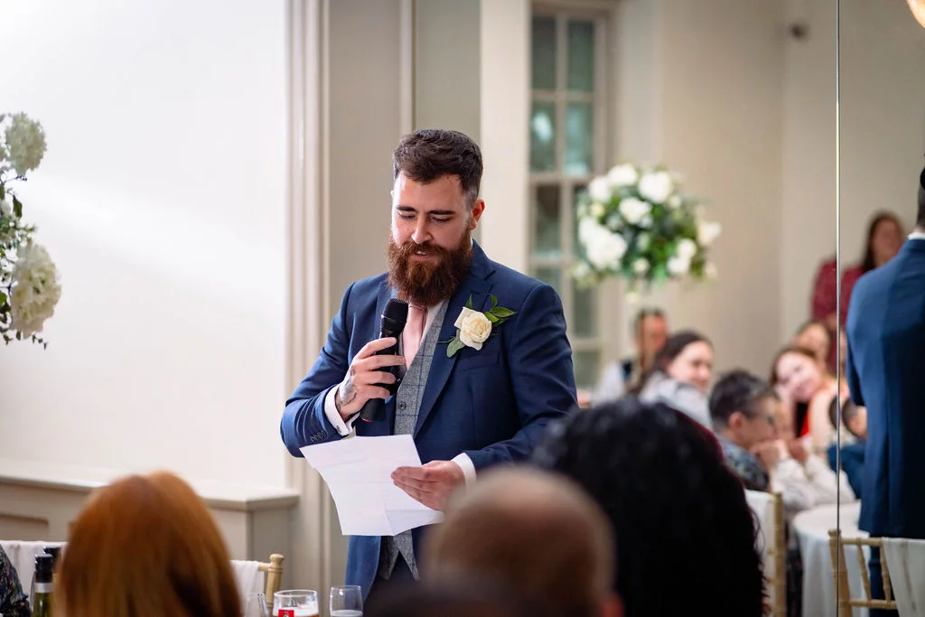 A bearded groom in a navy suit and tweed waistcoat delivers a heartfelt speech, reading from notes while holding a microphone at the wedding reception.