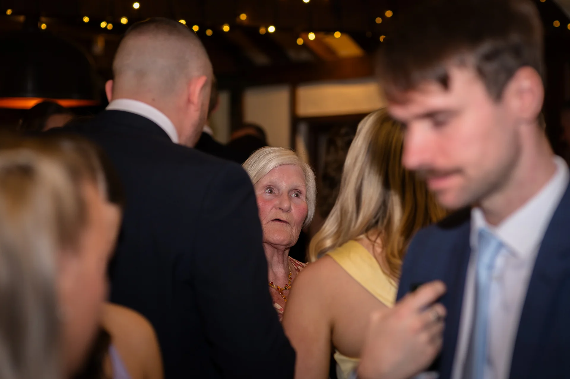 An elderly female guest looks on thoughtfully amidst a crowded evening wedding reception illuminated by overhead fairy lights.
