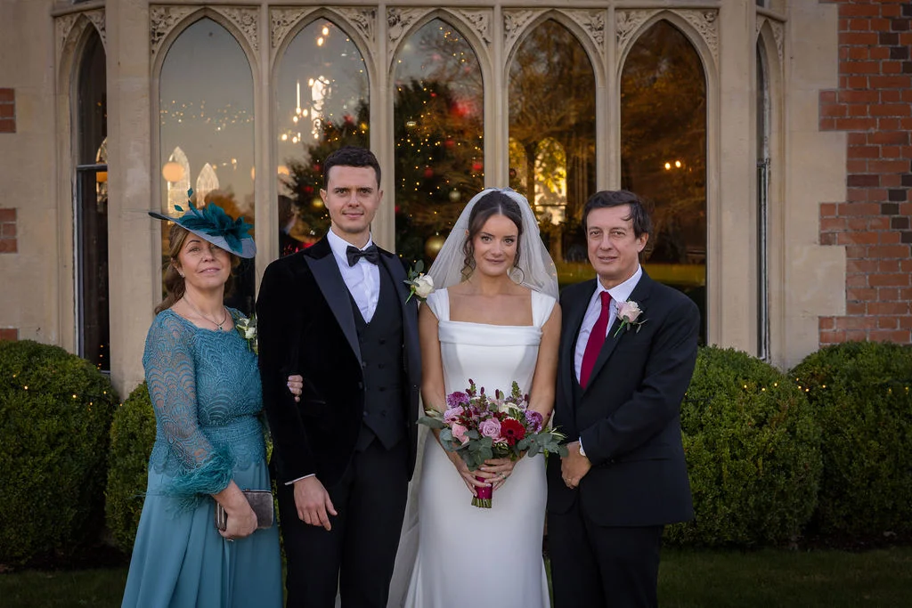A newlywed couple poses outdoors with their parents in front of a grand manor house window featuring a festive Christmas tree reflection.