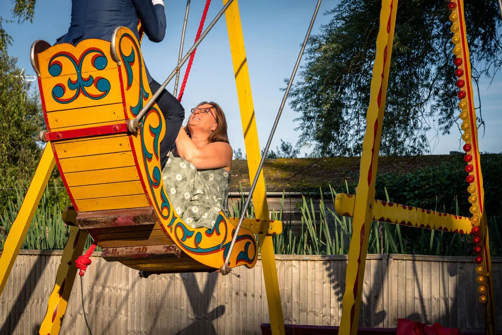 A wedding guest joyfully laughing while riding on a brightly painted vintage yellow and red funfair swing boat under a clear blue sky.