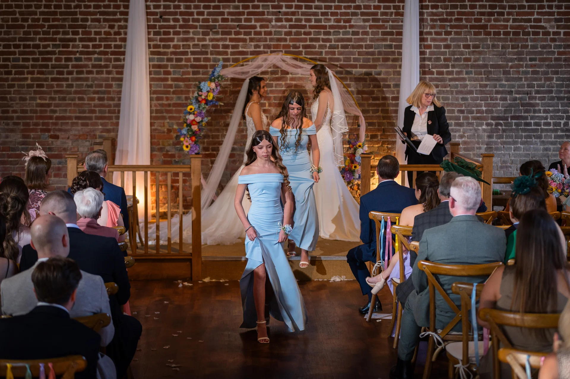 Bridesmaids walk down the aisle in dusty blue off-the-shoulder dresses during an indoor barn ceremony with an exposed brick backdrop and floral arch.