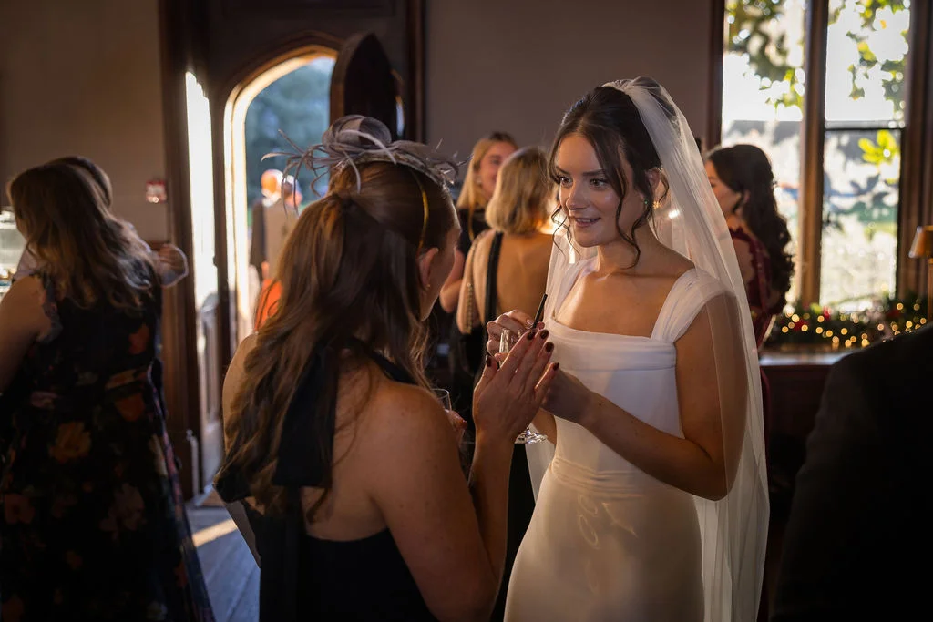 A smiling bride wearing a square-neck gown and veil chats with guests at a lively drinks reception inside a manor house.