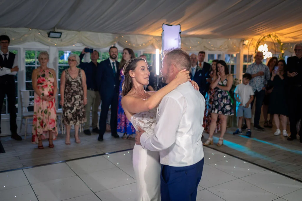 The newlywed couple share their romantic first dance on an illuminated LED dance floor inside a marquee surrounded by watching wedding guests.