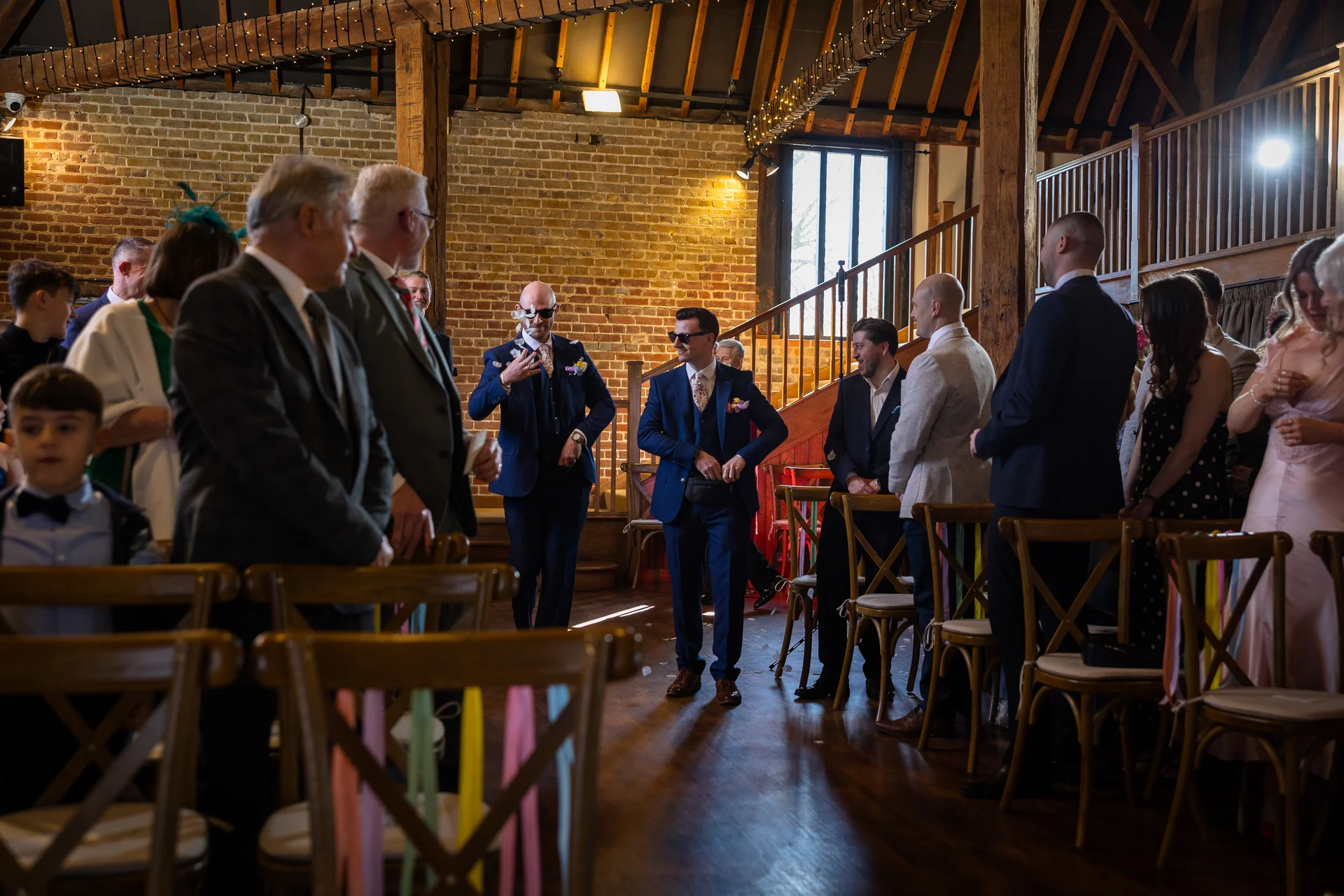 Guests and groomsmen gather inside a rustic barn wedding venue with exposed brick and wooden beams as grooms walk down the aisle wearing sunglasses.