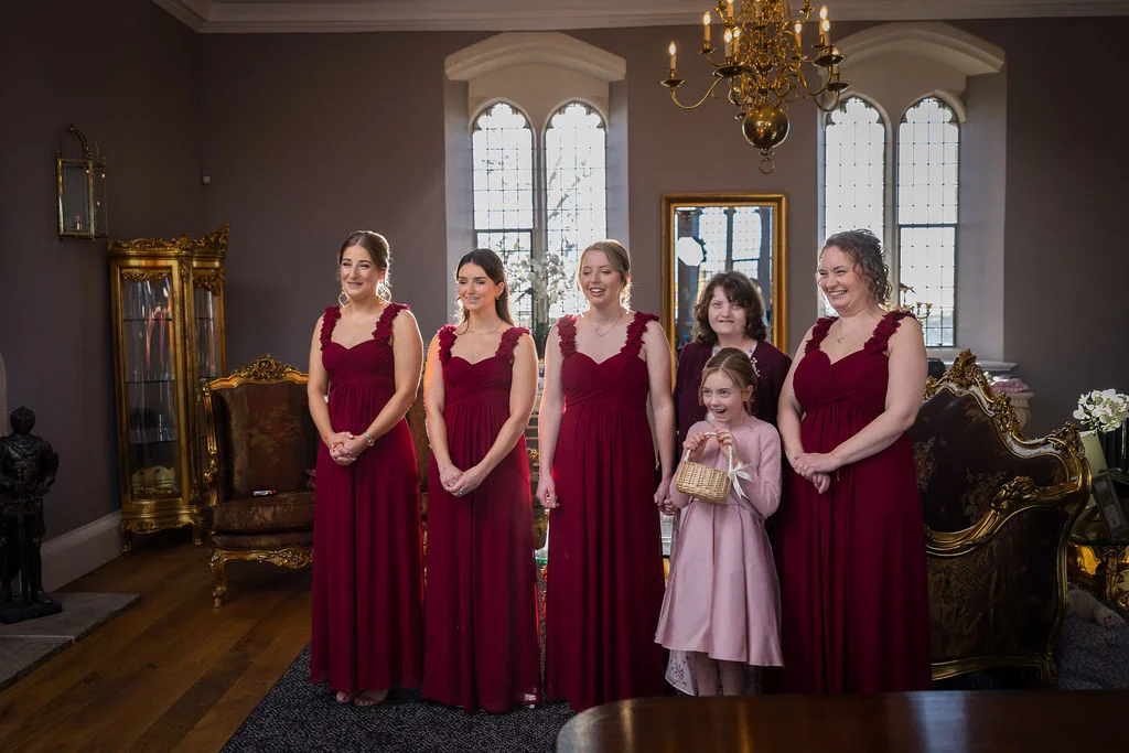 A group of bridesmaids wearing matching floor-length burgundy dresses pose together inside an elegant country house drawing room with tall arched windows.