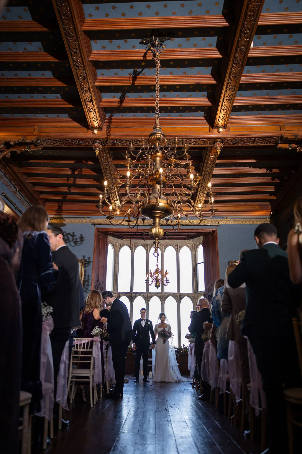 A newly married couple stands hand in hand at the end of the aisle during their castle wedding ceremony beneath a magnificent star-painted wooden ceiling.