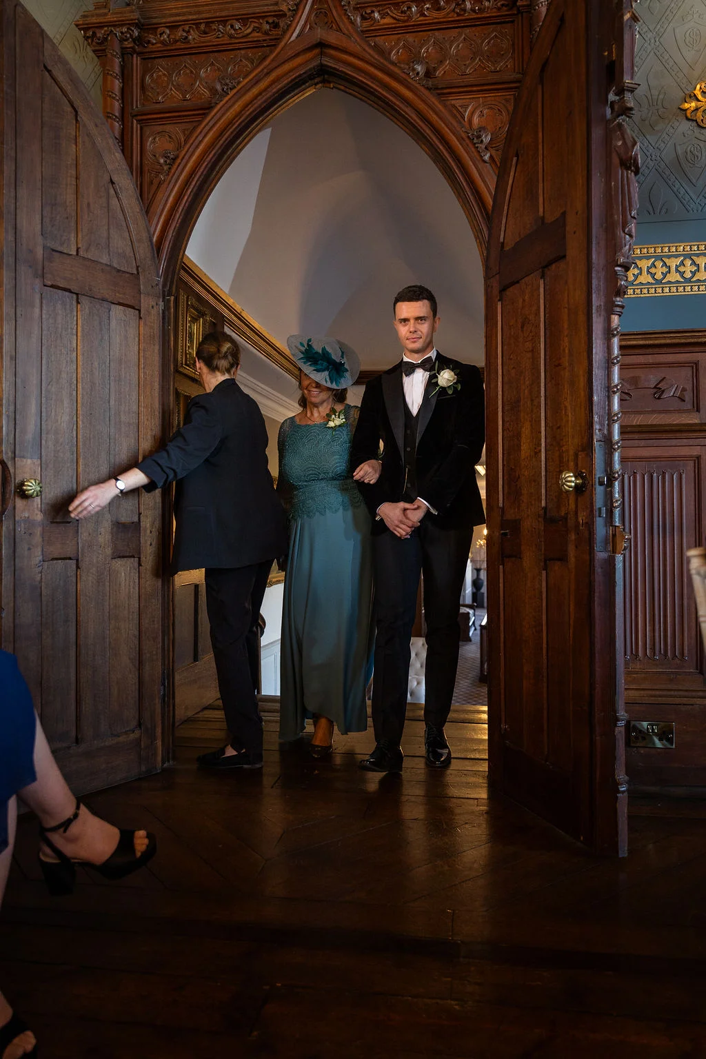 A groom in a black velvet tuxedo and a guest wearing a large fascinator enter through grand wooden doors into a panelled room.