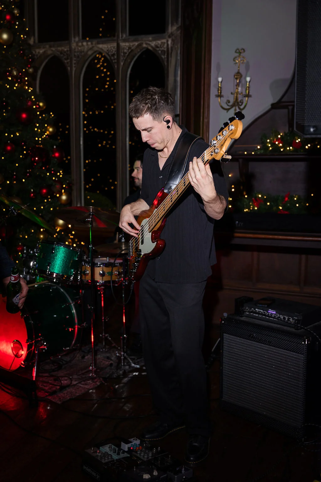A bassist performing on stage with his band at a festive Christmas wedding reception, with a decorated tree visible in the background.