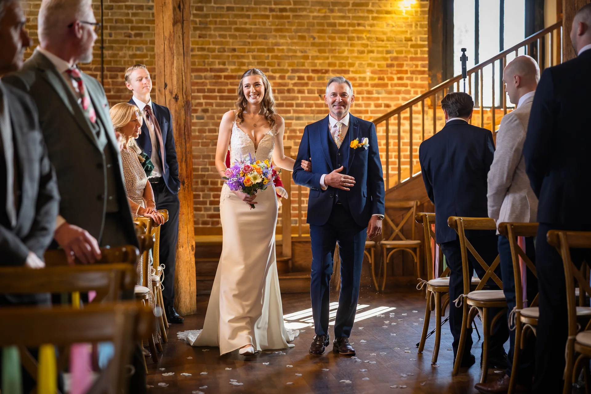 A smiling bride walks down the aisle arm in arm with her father in a rustic barn venue featuring exposed brick walls and wooden chairs.