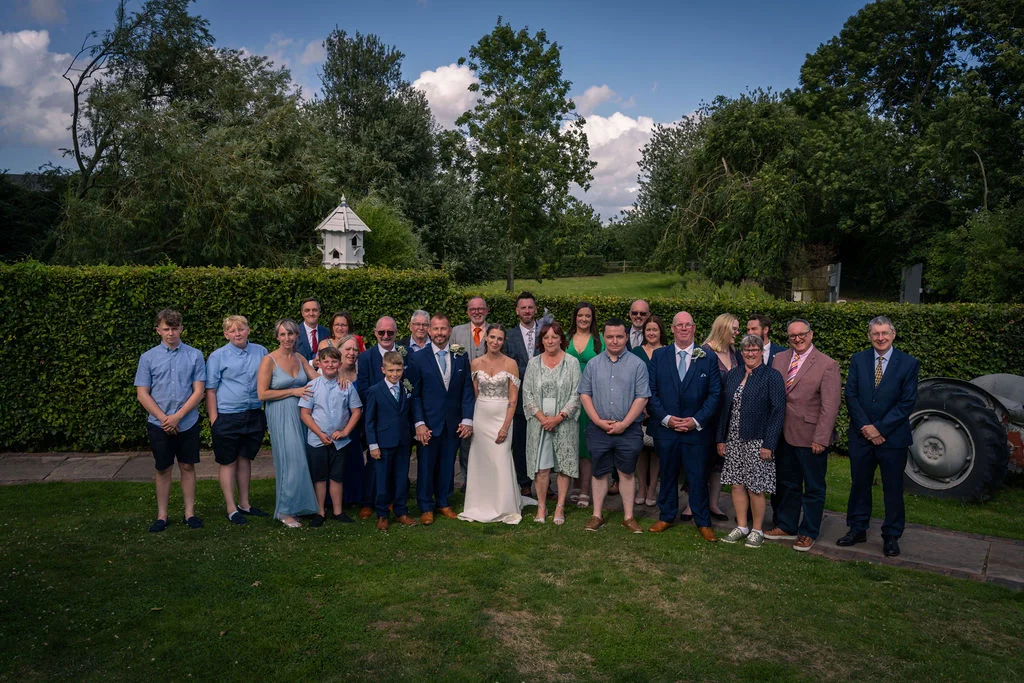 A large wedding party and family group pose together outdoors on a manicured lawn in front of a tall green hedge under a bright blue sky on a sunny summer day.