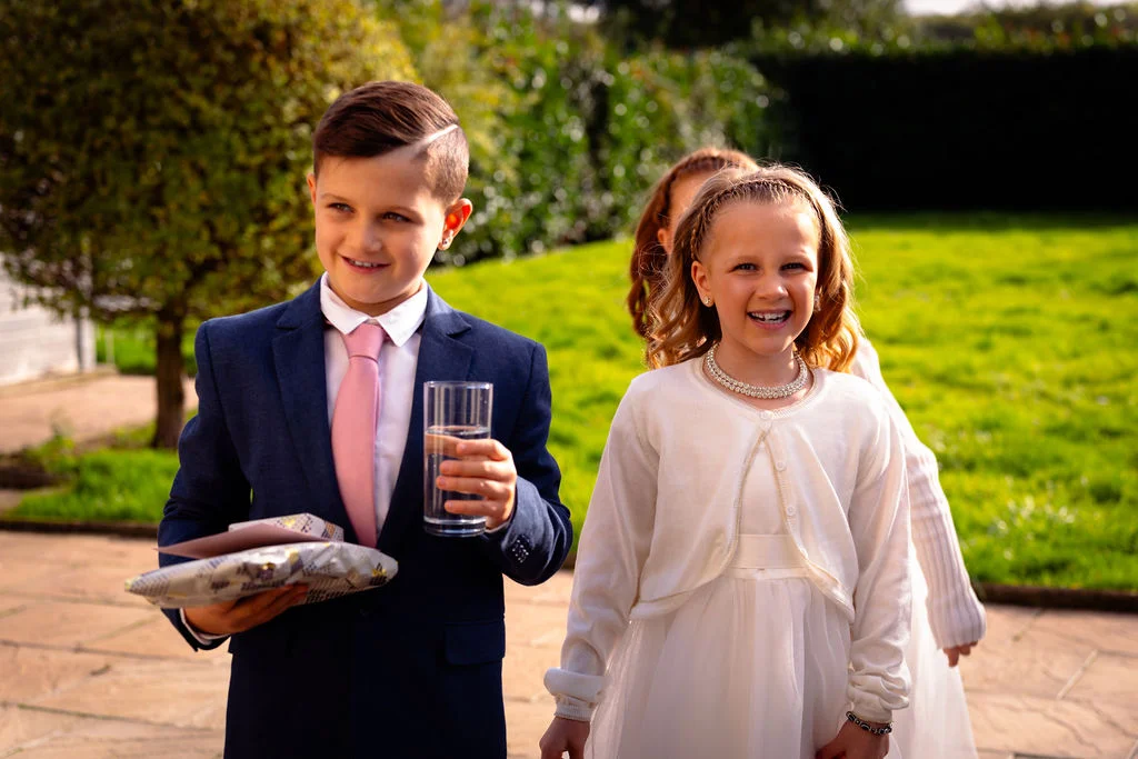 A young page boy in a navy suit with a pink tie holding a glass of water and a gift box walks outdoors alongside smiling young flower girls in white dresses during a sunny garden wedding.